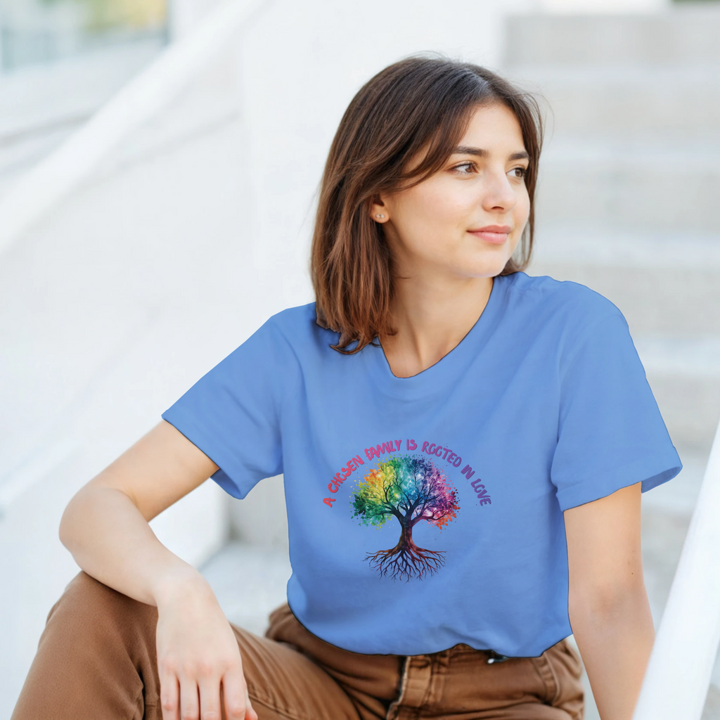 A woman wearing a Carolina Blue t-shirt featuring a colorful graphic of a tree with a rainbow-colored canopy and roots, with the text 'A Chosen Family is Rooted in Love' arched above it in pink and purple lettering.