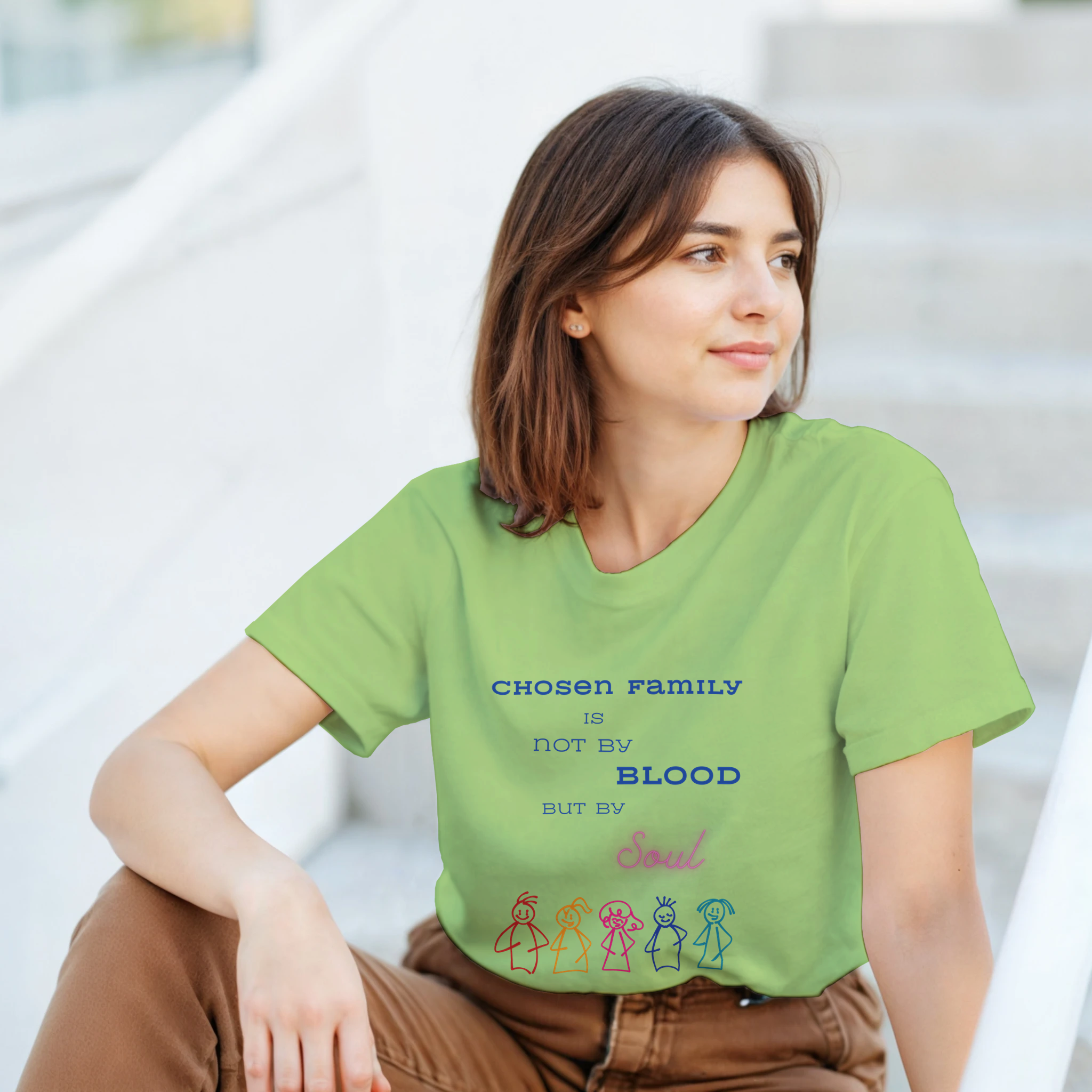 A women wearing a kiwi colored green t-shirt featuring a printed graphic with the text 'CHOSEN FAMILY IS NOT BY BLOOD BUT BY Soul' in blue and pink lettering, above five colorful hand-drawn stick figures.