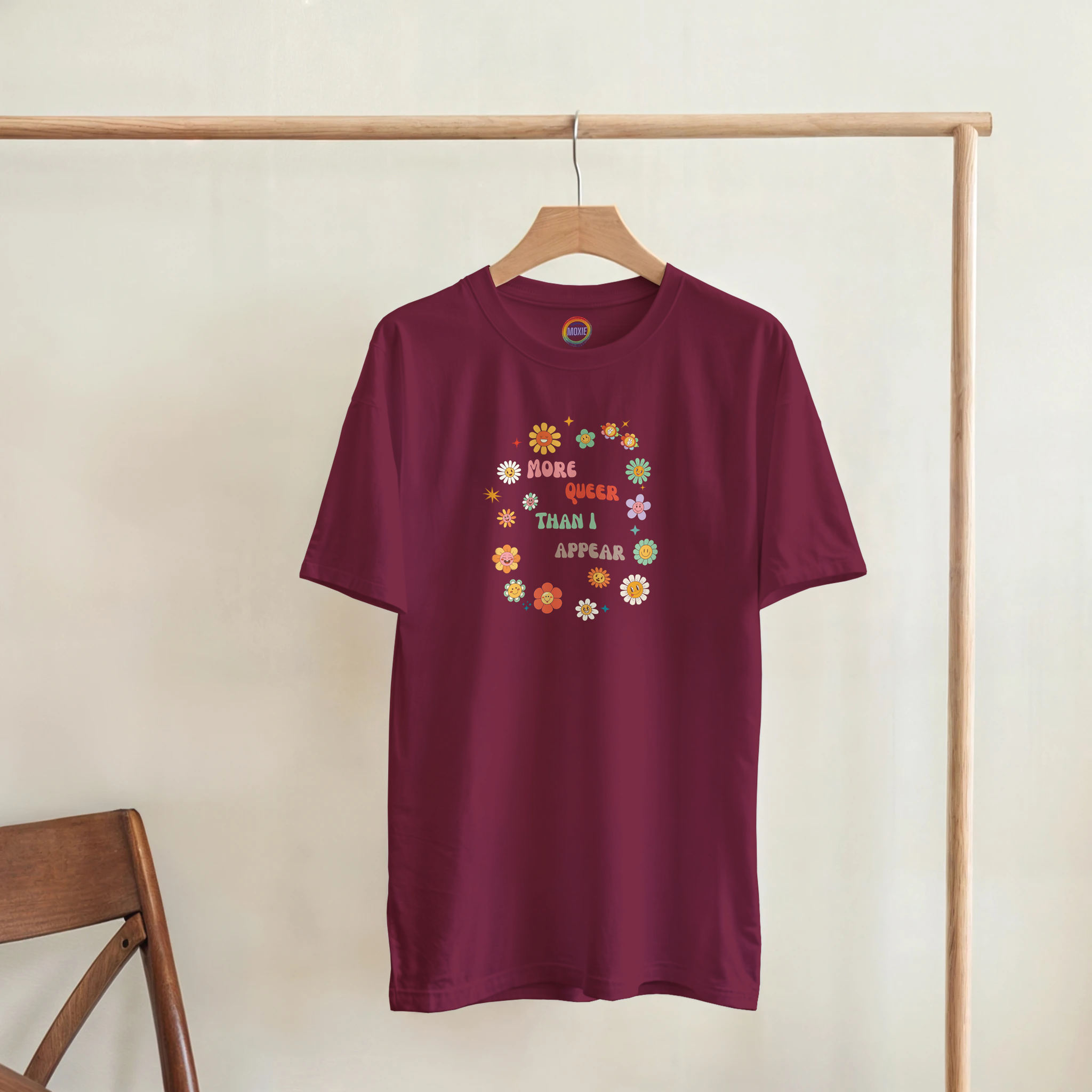 A maroon short-sleeve t-shirt hanging on a wooden hanger from a clothing rack, featuring a colorful retro-style graphic with flowers and the text 'More queer than I appear'.
