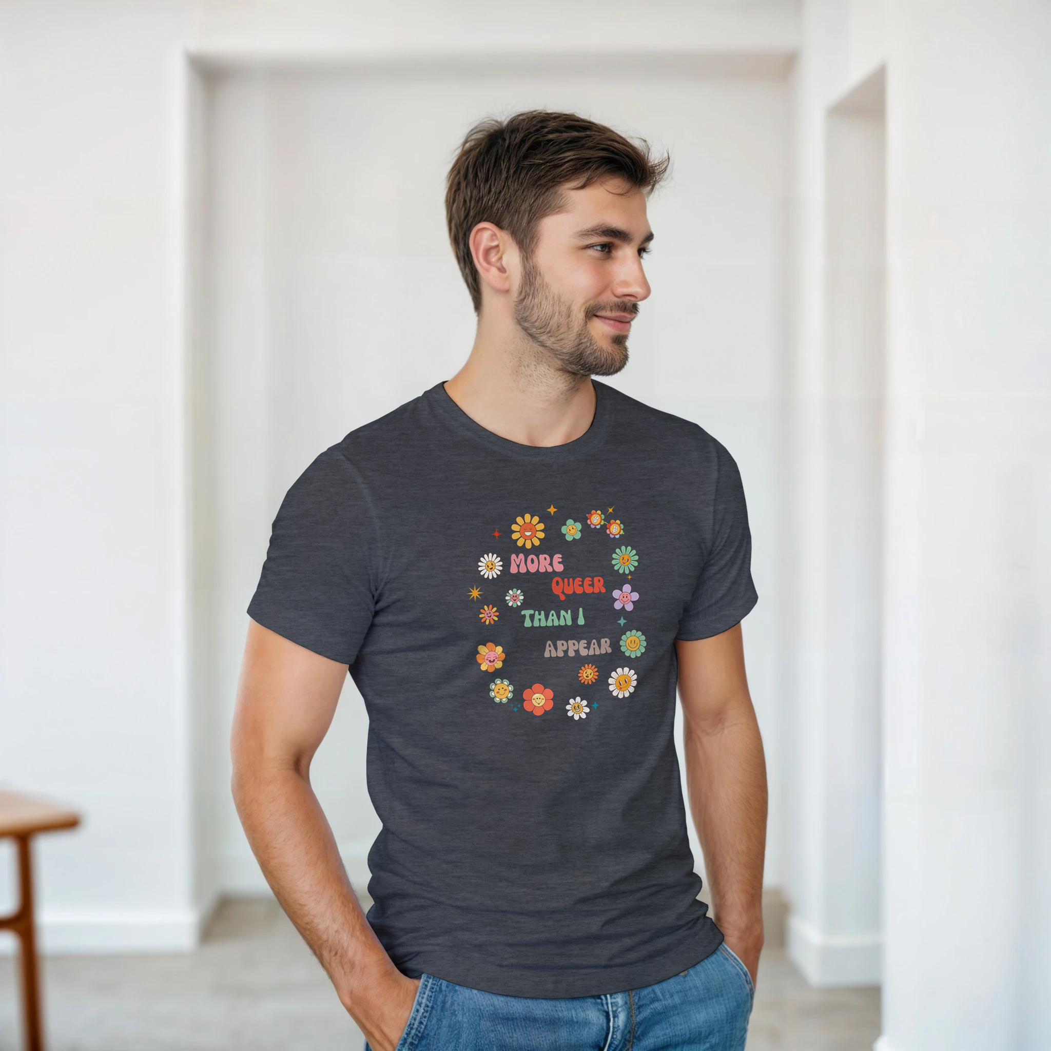 A dark heather tee with a graphic design featuring the text 'More queer than I appear' in a playful, retro font, surrounded by a circle of smiling, colorful flowers.