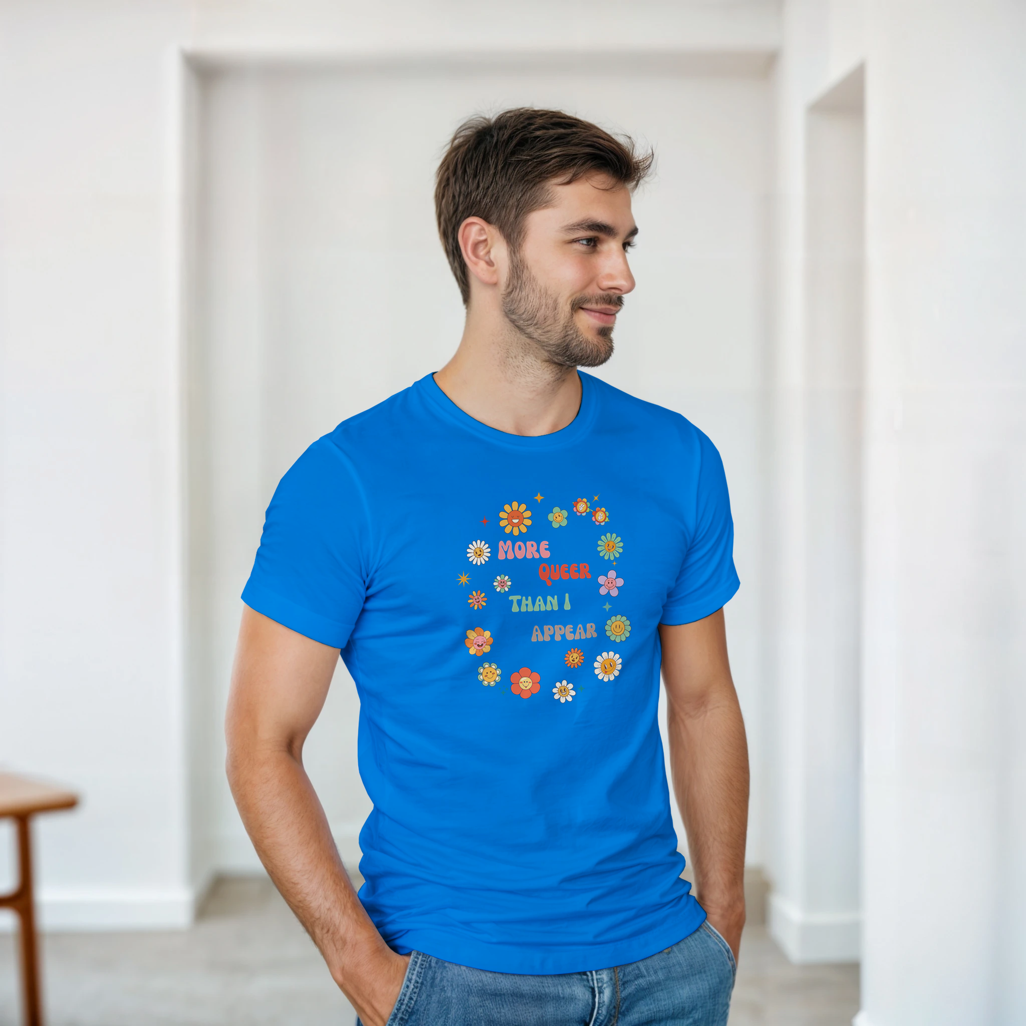 A man wearing a royal blue tee with a graphic design featuring the text 'More queer than I appear' in a playful, retro font, surrounded by a circle of smiling, colorful flowers.