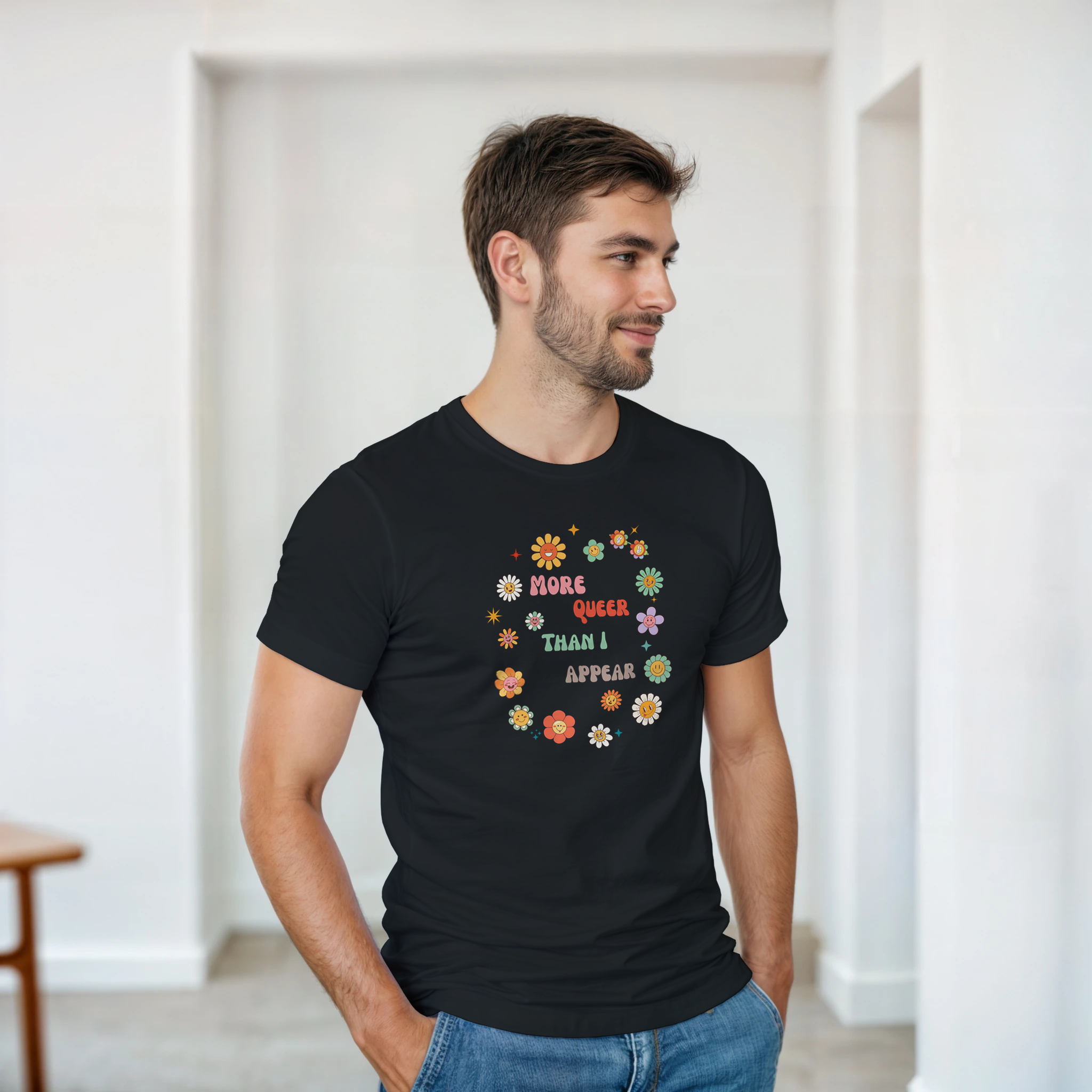 A black tee with a graphic design featuring the text 'More queer than I appear' in a playful, retro font, surrounded by a circle of smiling, colorful flowers.
