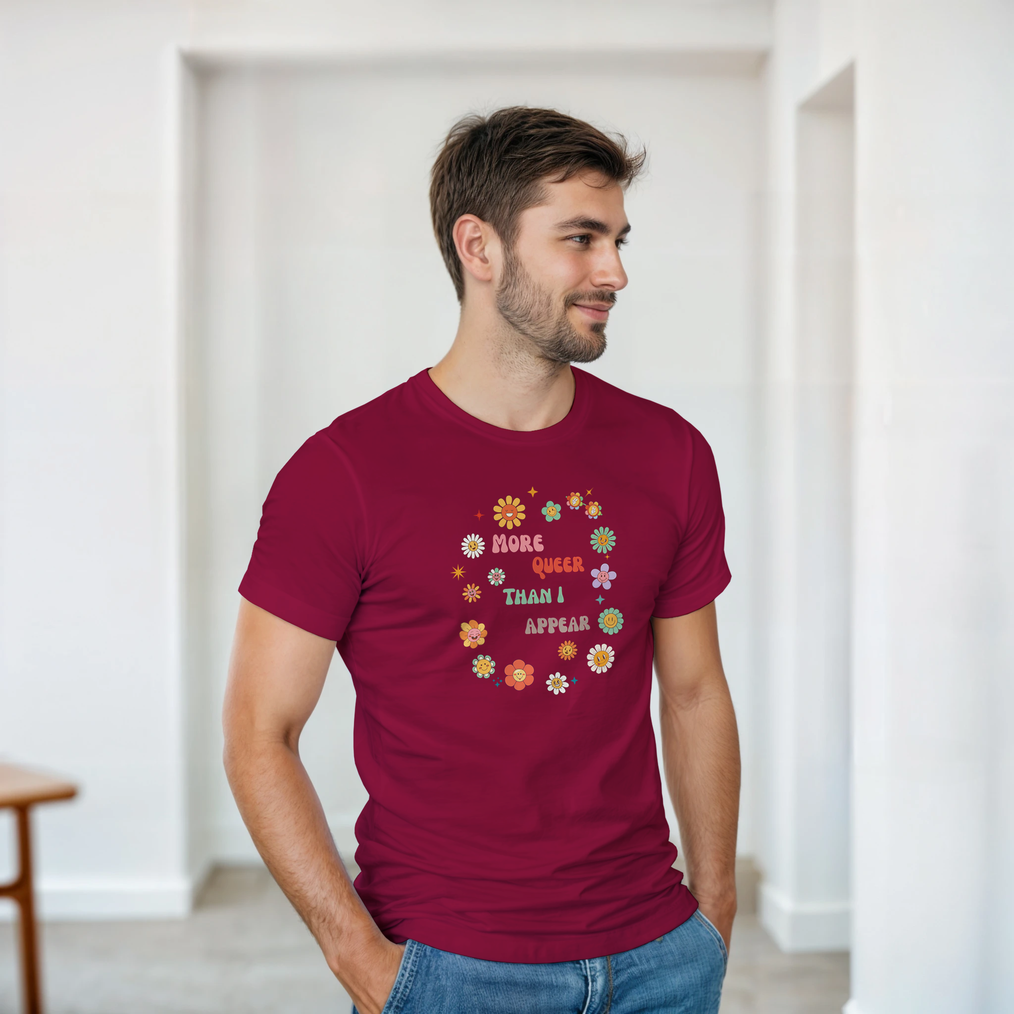 A maroon tee with a graphic design featuring the text 'More queer than I appear' in a playful, retro font, surrounded by a circle of smiling, colorful flowers.