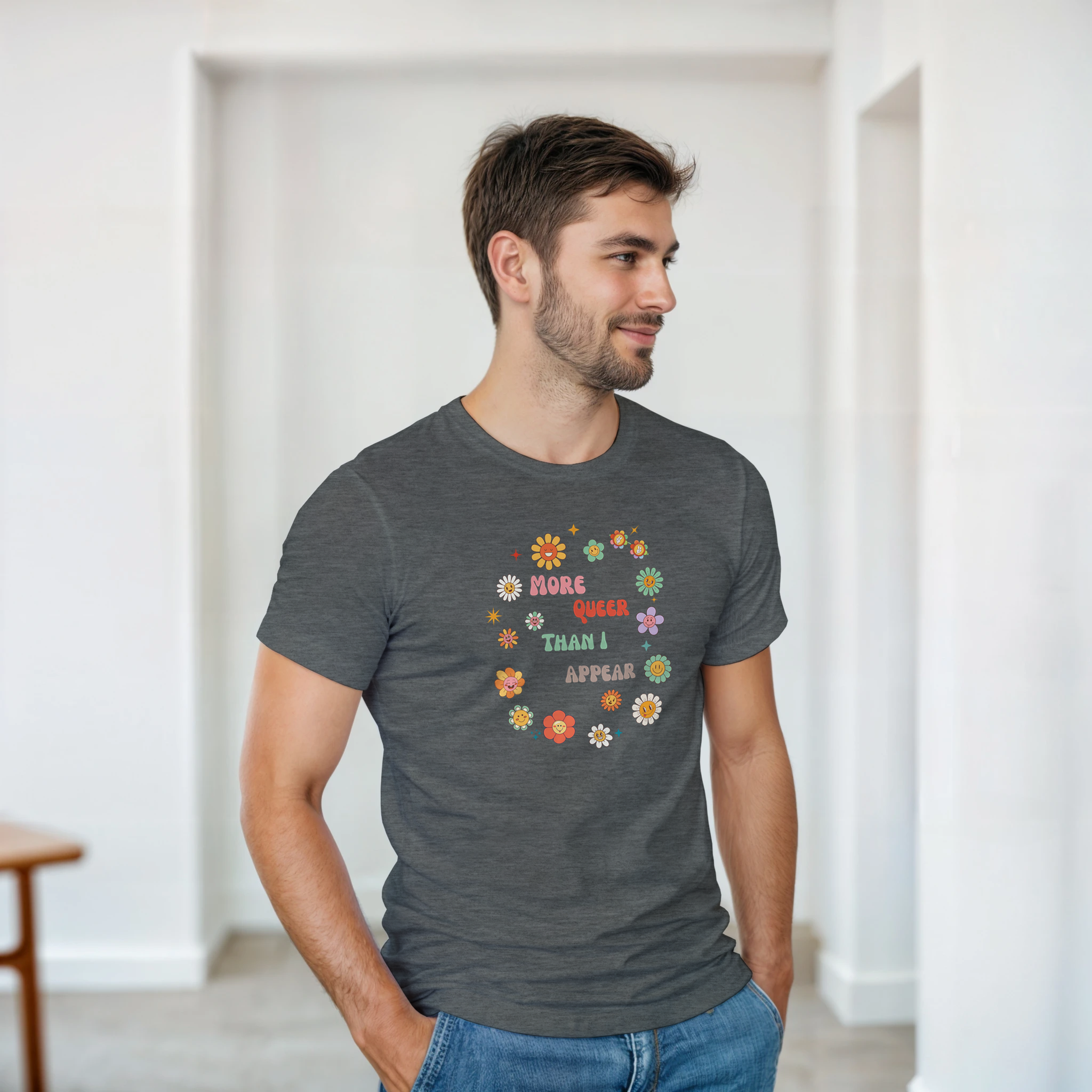 A dark heather tee with a graphic design featuring the text 'More queer than I appear' in a playful, retro font, surrounded by a circle of smiling, colorful flowers.