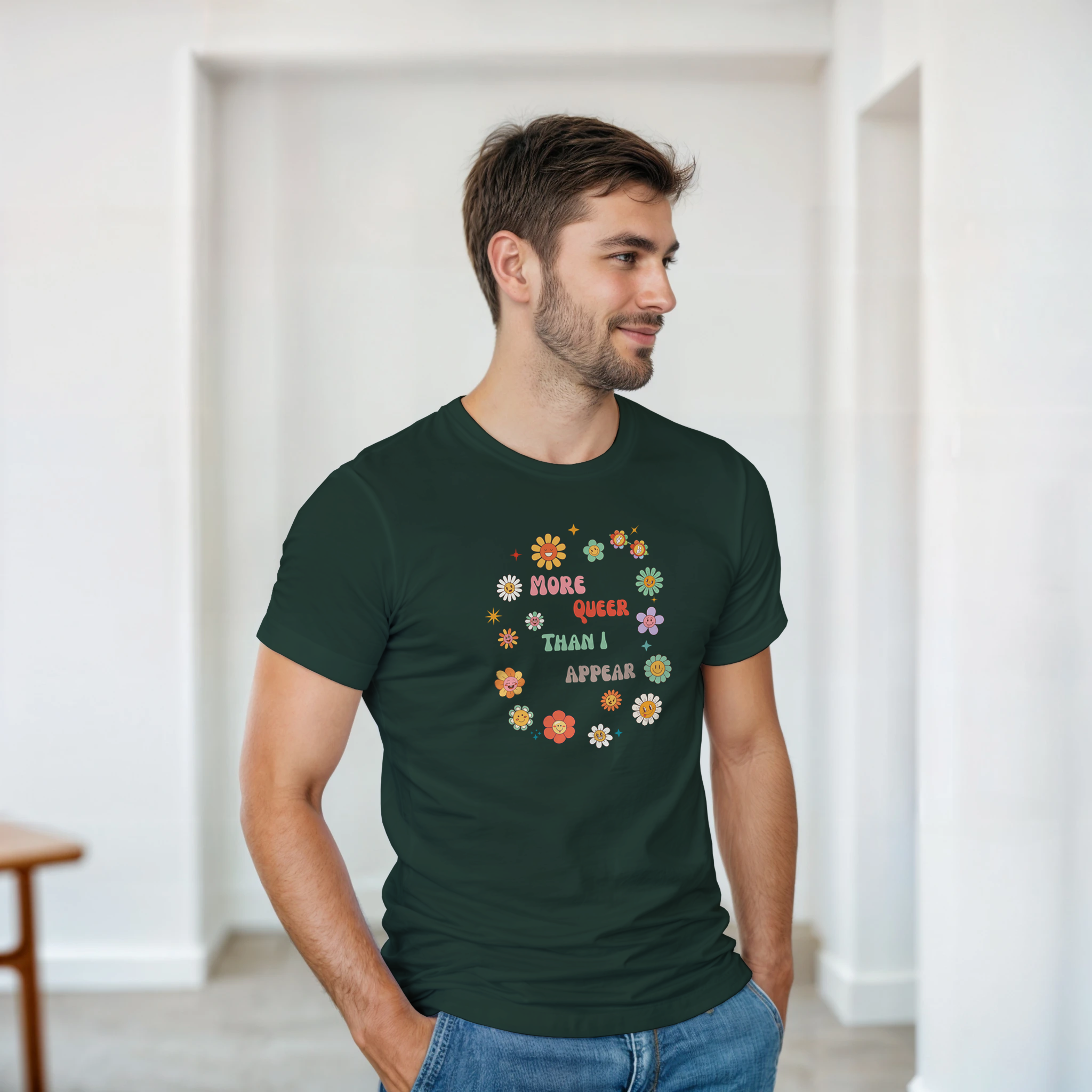 A forest green tee with a graphic design featuring the text 'More queer than I appear' in a playful, retro font, surrounded by a circle of smiling, colorful flowers.