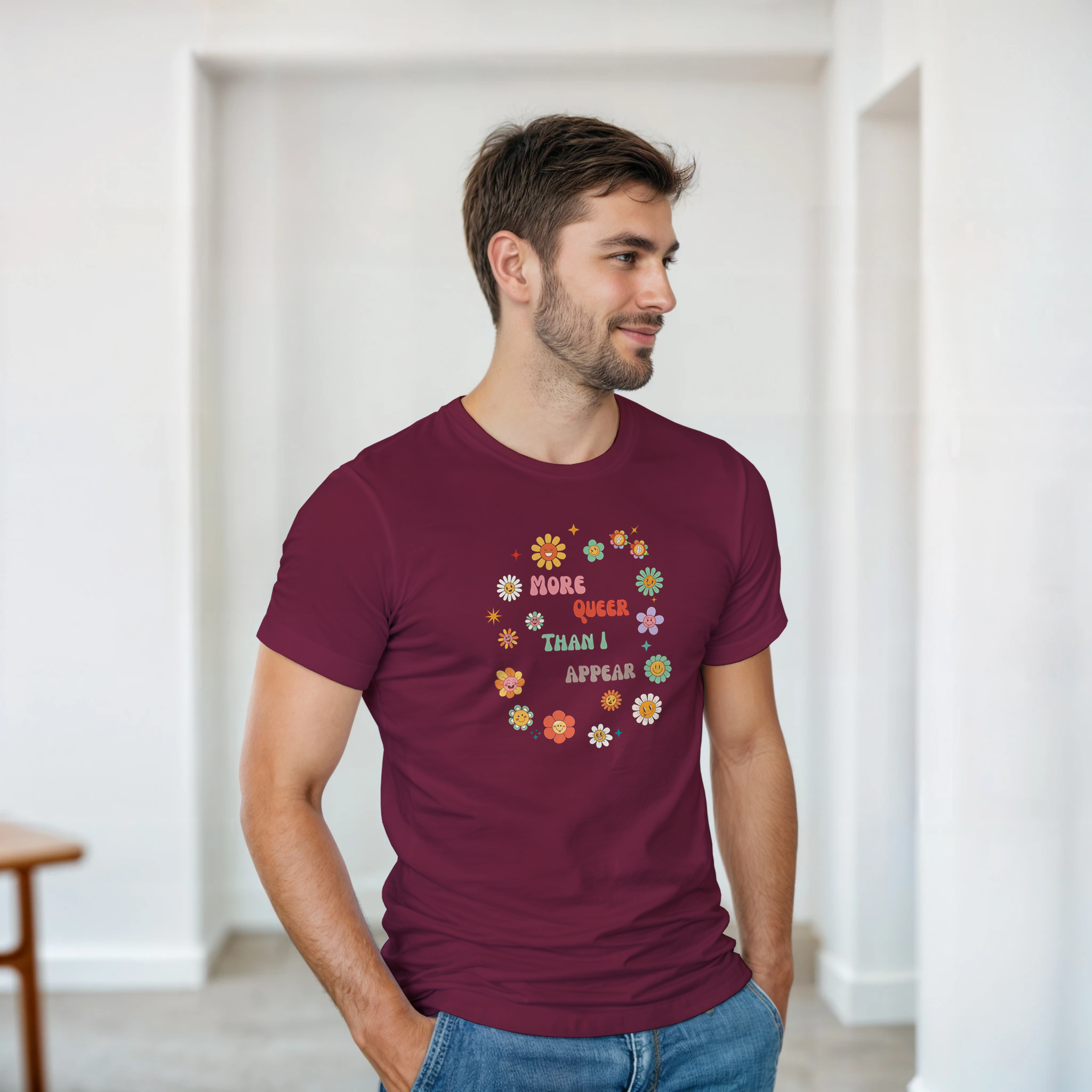 A maroon tee with a graphic design featuring the text 'More queer than I appear' in a playful, retro font, surrounded by a circle of smiling, colorful flowers.
