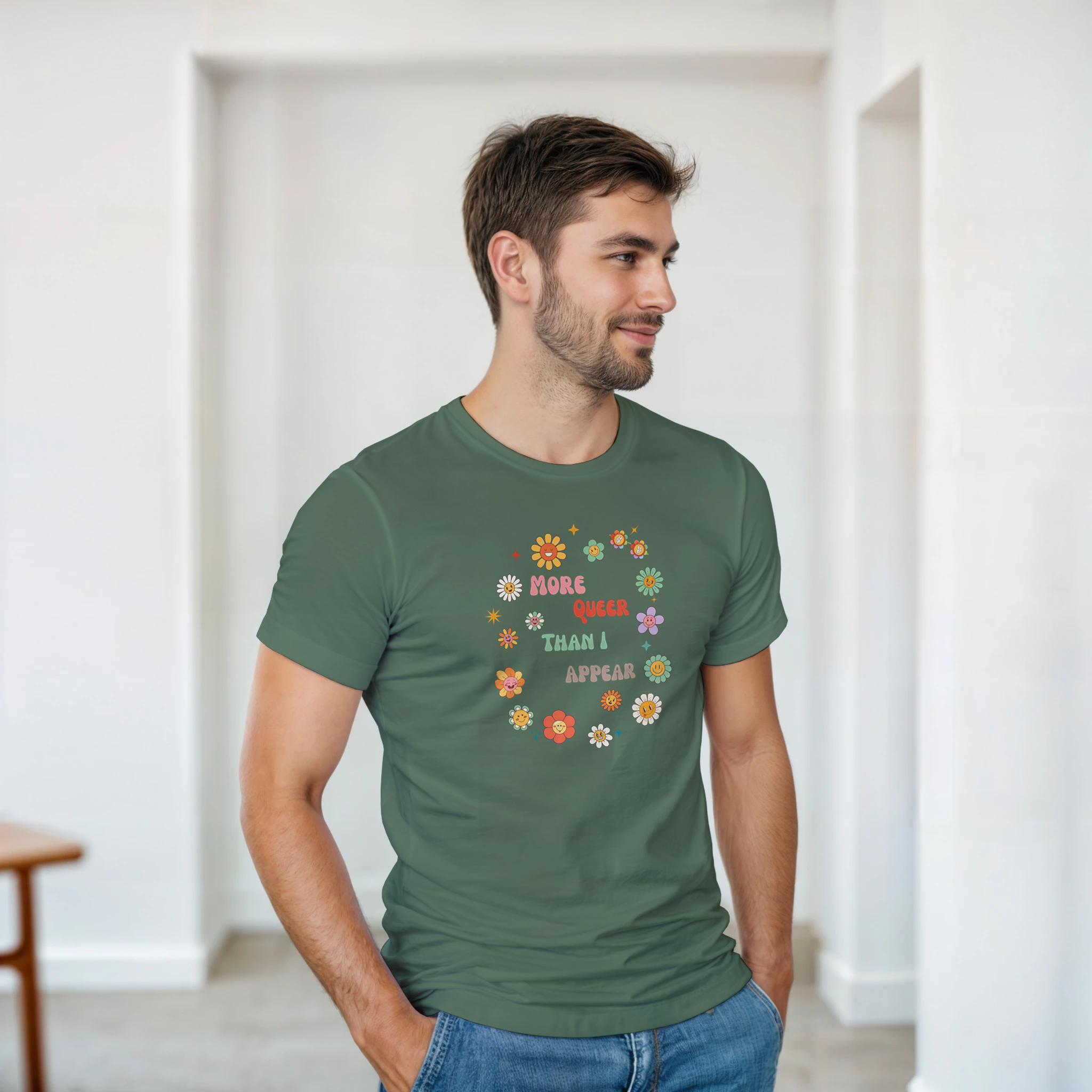 A man wearing a military green tee with a graphic design featuring the text 'More queer than I appear' in a playful, retro font, surrounded by a circle of smiling, colorful flowers.