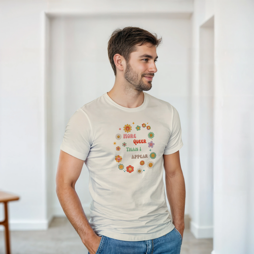 A natural beige tee with a graphic design featuring the text 'More queer than I appear' in a playful, retro font, surrounded by a circle of smiling, colorful flowers.