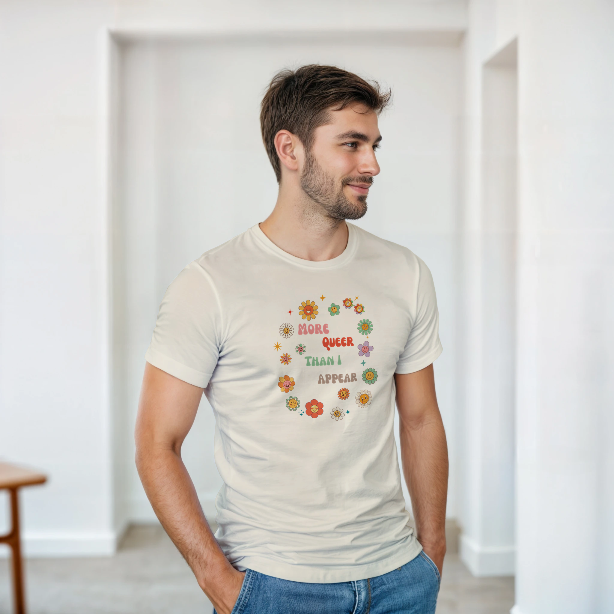 A natural beige tee with a graphic design featuring the text 'More queer than I appear' in a playful, retro font, surrounded by a circle of smiling, colorful flowers.