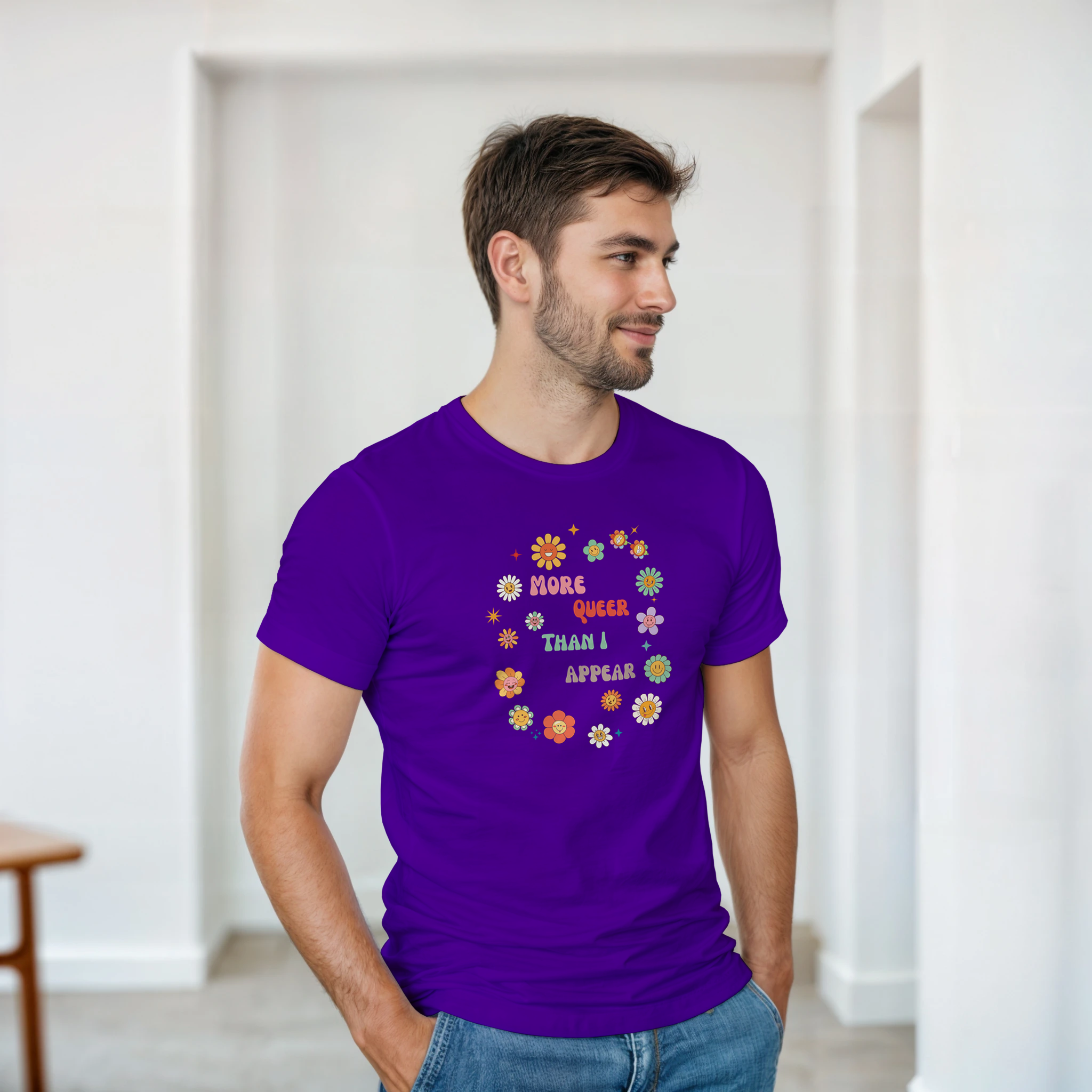 A purple tee with a graphic design featuring the text 'More queer than I appear' in a playful, retro font, surrounded by a circle of smiling, colorful flowers.