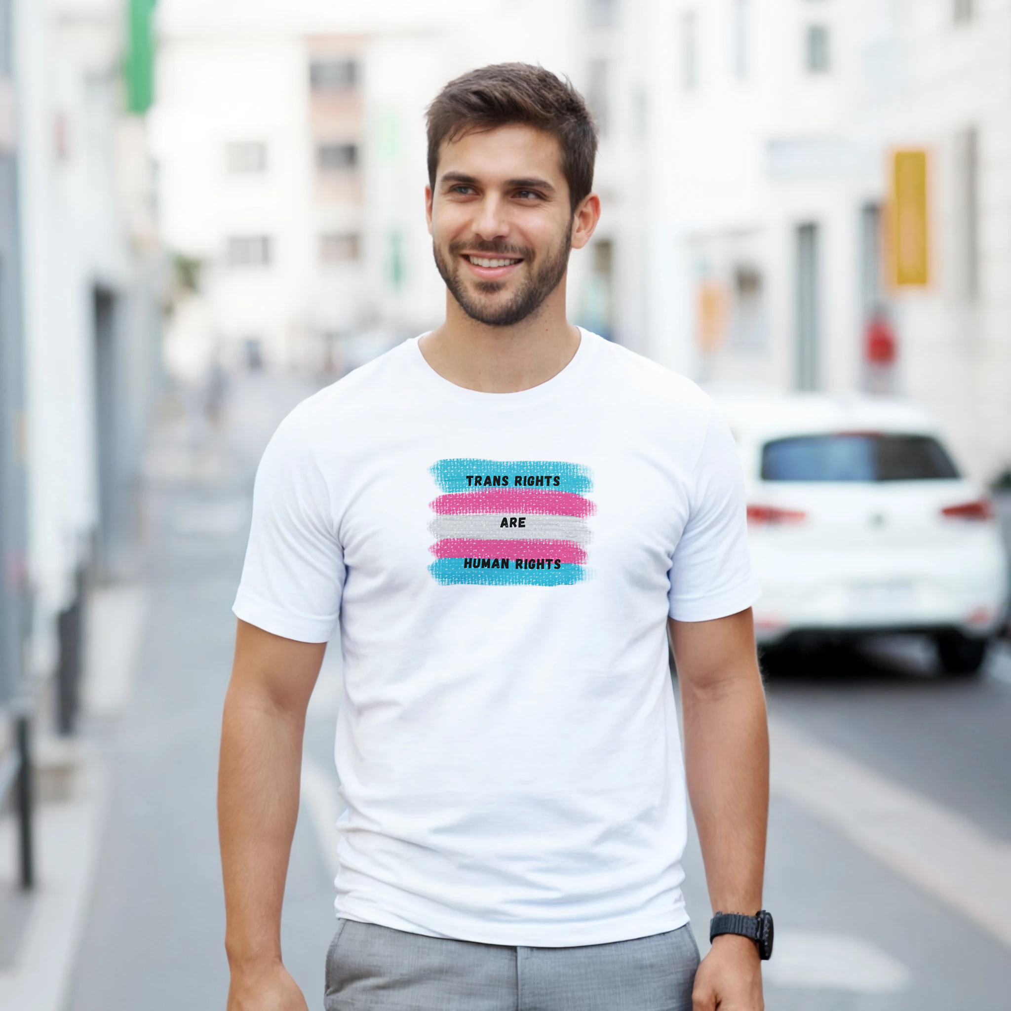 A close-up of a white t-shirt featuring a printed graphic of the transgender pride flag with the text 'TRANS RIGHTS ARE HUMAN RIGHTS' in black capital letters. The back ground is swatches showing trans flag colors.