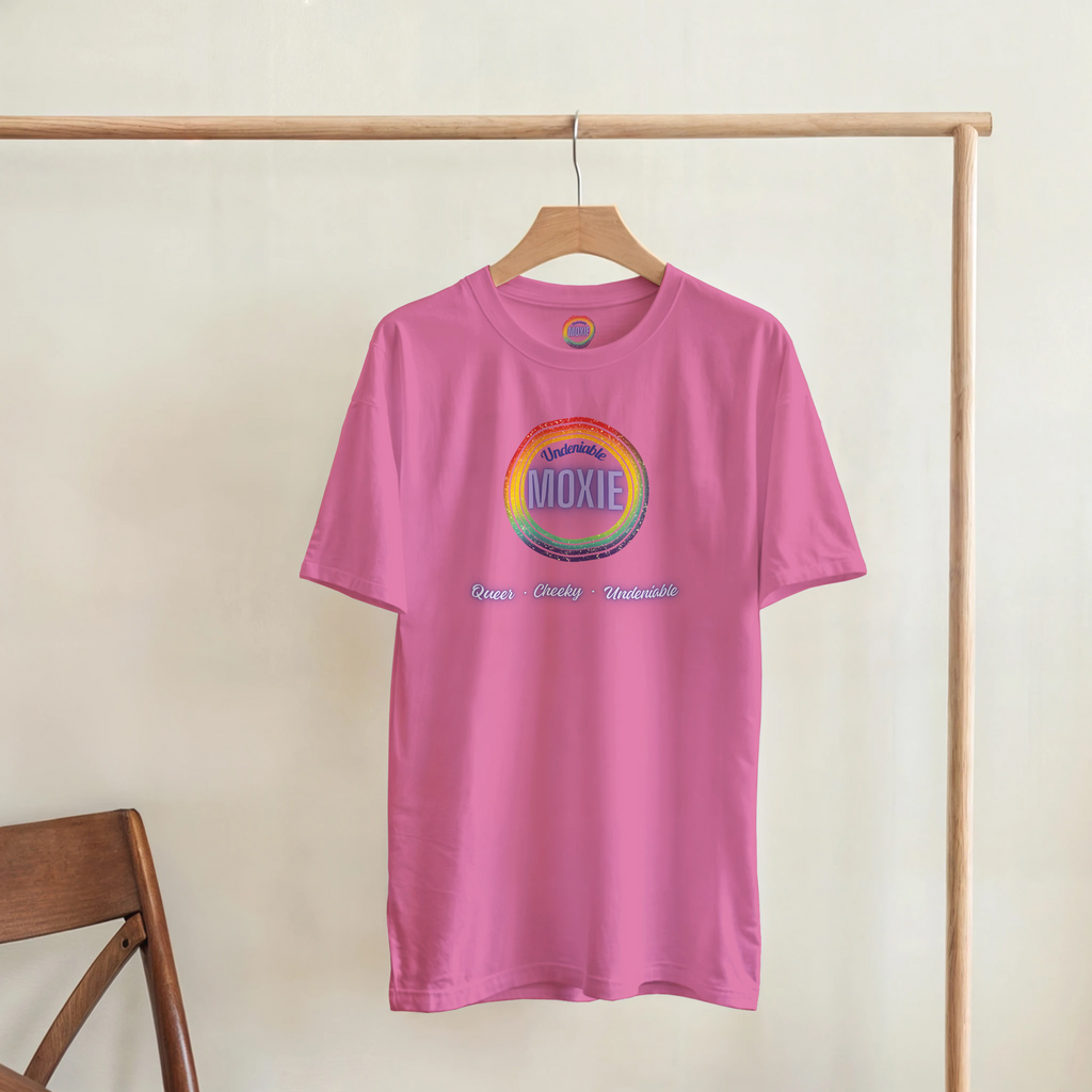 Graphic print on an azalea colored t-shirt hanging on a rack featuring a circular rainbow design with the text 'Undeniable MOXIE' and 'Queer • Cheeky • Undeniable' written below in a stylized font.